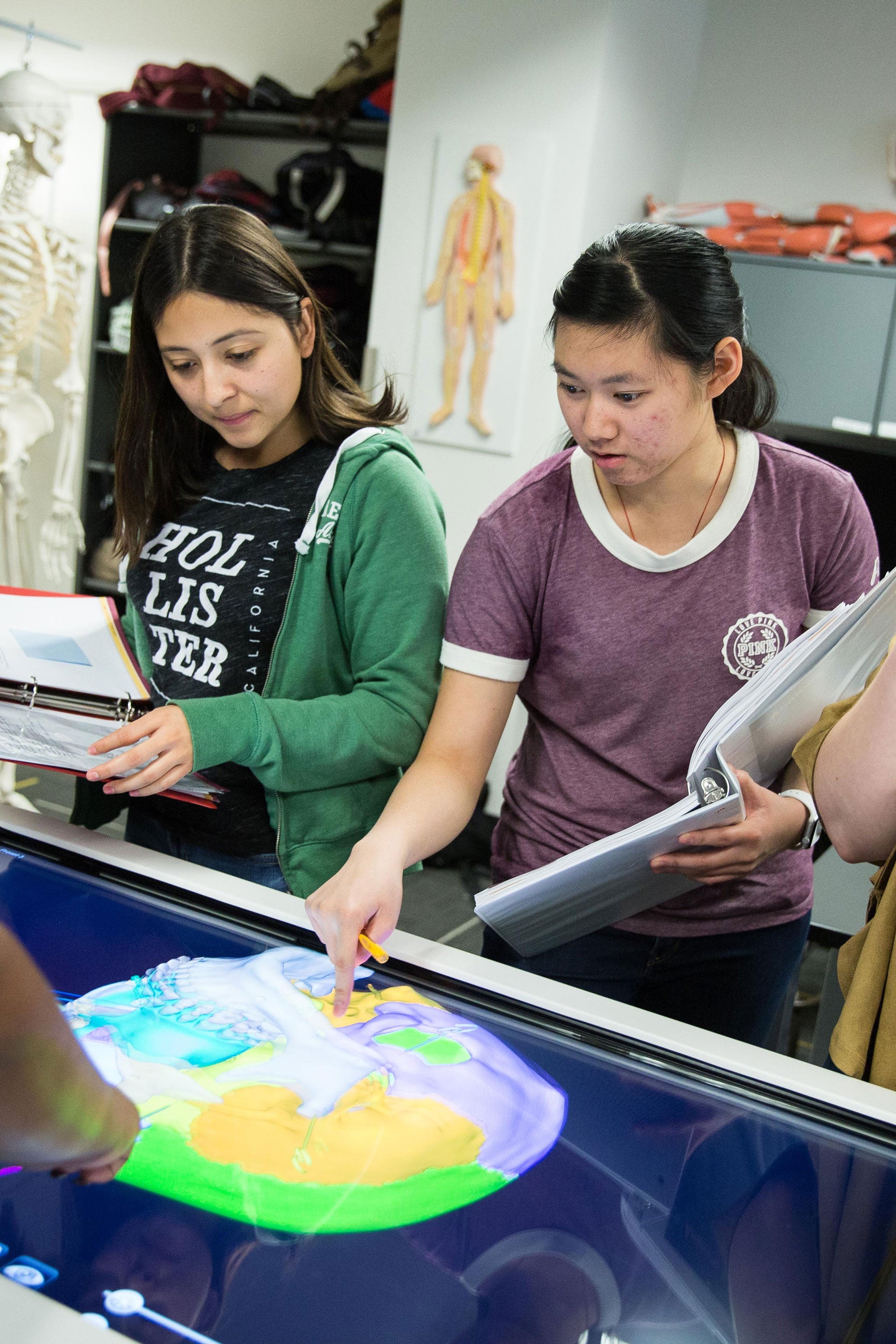 two young women standing in front of a digital table holding notebooks