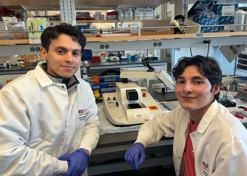 Two male students with dark hair sitting in at a table with lab equipment behind them